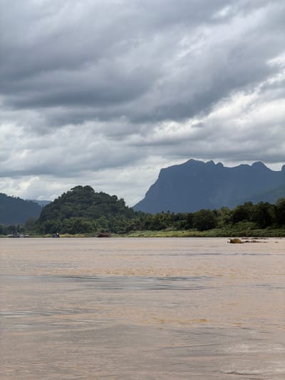 The image shows a wide, muddy river with a heavily overcast sky. In the background, dense green foliage covers rolling hills and dramatic, jagged mountains. Several boats are visible on the river and along the shore, suggesting human activity and possibly a settlement. The overall impression is one of a natural landscape under a somber sky. The specific location is the Mekong River near Siao, Laos.