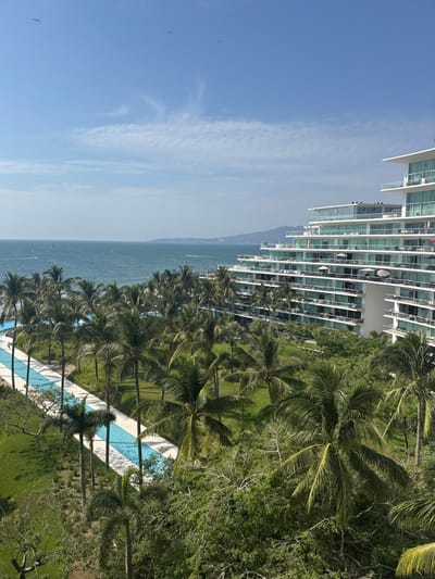 A high-rise building stands beside a tropical ocean view. Palm trees line the shore, leading to a long, narrow swimming pool. The blue sky contains faint clouds. The building's modern architecture features stacked balconies. The sea is calm, with hints of distant land. Lush greenery surrounds the pool area and extends toward the building.