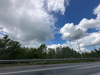 The image captures a roadside scene with a clear blue sky filled with dramatic white and gray clouds. A dark asphalt road with a white line runs diagonally across the foreground, suggesting movement or travel. To the left and center, a dense line of dark green trees forms a natural backdrop, with some taller trees interspersed. A silver guardrail runs horizontally across the middle of the image, separating the road from the grassy verge and the trees beyond.

Dominating the upper right portion of the sky is a tall, slender lamppost, leaning slightly as if angled towards the viewer. Further to the right, a few utility poles with wires are visible amidst the trees. The contrast between the bright, fluffy clouds and the darker, more overcast ones hints at dynamic weather conditions, perhaps a day with passing showers or rapidly changing skies. The greenery suggests a temperate season, likely summer or late spring. The overall atmosphere is one of being on a journey through a rural or semi-rural landscape under a vast, active sky. No people or text are visible in the image. The setting is consistent with a road in Chernovskii, Russia.