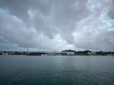 The image captures a wide-angle view of a harbor or bay with a city skyline in the background, under an overcast sky. The foreground is dominated by dark blue, choppy water. In the mid-ground, a coastline stretches horizontally across the frame. Prominently featured on the coastline is a modern, white, lattice-like structure, likely a stadium or convention center. To the left of this structure, several buildings of varying architectural styles, including a tall white building and a building with a spire, are visible, suggesting an urban setting. A strip of green trees separates the city buildings from the water. Several boats of different sizes are moored or moving in the water. The sky above is filled with heavy, grey clouds, with some patches of lighter sky peeking through, indicating a potentially stormy or cloudy day. The overall scene depicts a waterfront area of a city, likely Pointe-à-Pitre, France, given the architecture and waterfront setting. There are no visible people in the image.