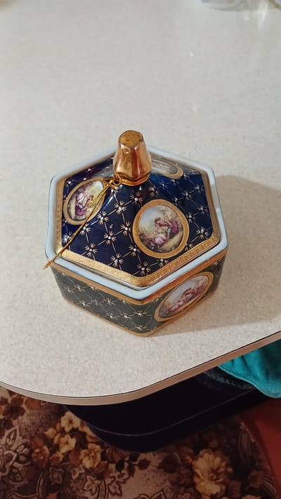 A hexagonal, dark blue ceramic box with gold trim and decorative paintings of people is placed on a light-colored, speckled table. The box has a gold, pointed finial on top, from which a thin gold cord hangs. The surface of the table is textured and appears to be a tabletop, possibly made of laminate or a similar material. The edge of the table is visible, with a curved profile and a thin gold trim. Beneath the table, a dark surface is visible, possibly the underside of a shelf or a drawer, with a floral patterned fabric in brown and beige tones. A teal colored cloth is also visible peeking out from beneath the table. The image does not provide any information about the city of Yalama, Azerbaijan.