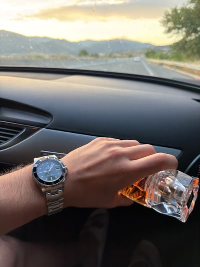 A right hand is holding a clear, multifaceted bottle containing amber liquid, inside a car. A wristwatch with a silver band and black face is on the wrist of the hand. The interior of the car is black. A blurred view of a landscape with greenery and sunlight is seen through the car window. The location is not discernible as Sidirokastro, Greece.