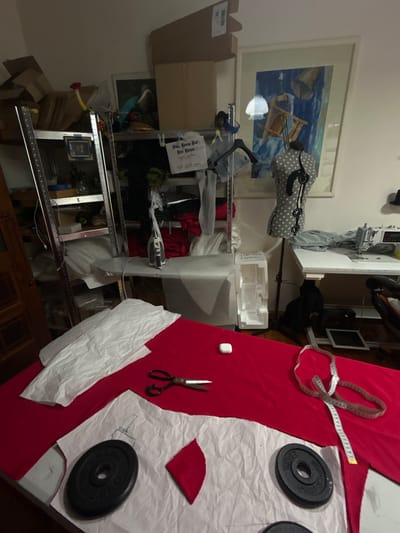 The image depicts a cluttered indoor scene. A metal shelving unit is filled with boxes, clothes, and other miscellaneous items. A sewing machine is partially visible behind the shelving unit. A framed painting hangs on the wall behind the shelving unit.

To the right of the shelving unit, a red fabric is spread across a surface, possibly a table. On the red fabric, there are white paper patterns, two black weight plates, a red scrap of fabric, a white button, a measuring tape, and a pair of scissors.

There is no indication of the image's location being Gryfino, Poland. No people are visible.