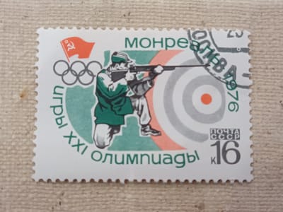 This is a photograph of a postage stamp. The stamp features a graphic illustration of a male athlete participating in a shooting competition, likely at the Olympic Games. The athlete is depicted in profile, wearing a green tracksuit and a green cap, aiming a rifle. In the background, there is a target with concentric circles, with the bullseye highlighted in orange. To the left of the athlete, the Olympic rings are shown, and above them, a red flag with a hammer and sickle symbol.

The stamp includes text in Russian and numerals. Along the bottom arc of the stamp, it reads "XXI ОЛИМПИАДЫ," which translates to "XXI Olympiad." At the top, "МОНРЕАЛЬ 1976" is visible, indicating the location and year of the Montreal 1976 Olympic Games. In the bottom right corner, a "ПОЧТА СССР" (POST OF USSR) emblem is present above the value "16 K" (likely 16 kopecks). The overall design suggests a commemorative stamp issued by the Soviet Union for the 1976 Olympics. The background of the photograph is a textured, light brown surface, possibly a piece of fabric or burlap. The lighting suggests an indoor setting.