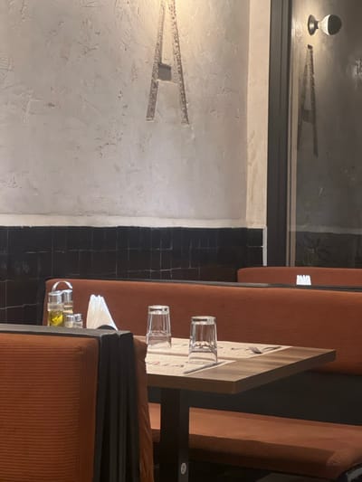 A restaurant's interior shows an orange booth seat with a table set for dining. Two glasses and condiments sit on the tabletop. The dark floor tiles contrast with a light-colored wall featuring an etched or raised horizontal design. A window near the ceiling reflects the interior lighting and design. The scene is captured from an overhead perspective.