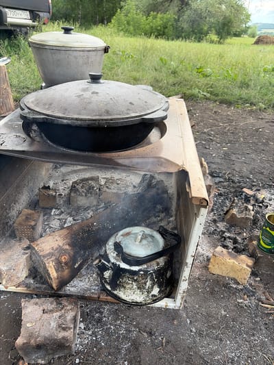 The image depicts an outdoor cooking setup in a rural setting, likely the village of Subkhankulovo, Russia, given the provided context. In the foreground, a makeshift stove constructed from metal and bricks is burning. Logs and ashes are visible within the stove, indicating an active fire. A well-used, blackened metal kettle is placed inside the stove, suggesting it is being heated.
On top of the stove, two large metal pots with lids are present. The larger pot in the background has a light-colored lid, while the pot in the foreground has a darker, presumably cast-iron lid. These appear to be traditional cooking vessels for outdoor preparation.
The surrounding environment consists of dry, dusty ground with scattered ashes and some larger pieces of brick. Behind the stove, tall, dry grass and green foliage are visible, with trees forming a backdrop. In the far background, a haystack and rolling hills suggest a rural or agricultural landscape. A vehicle is partially visible in the upper left corner of the image, with a license plate that appears to read "B700YH 702". The overall scene conveys a sense of rustic outdoor cooking and self-sufficiency. There are no people visible in the image.