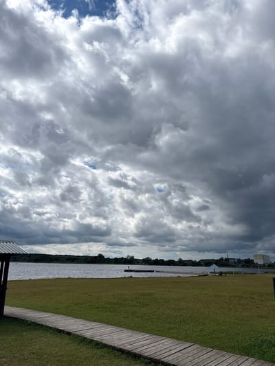The image depicts a cloudy day at a lakeside park in Mazalawski Sel'ski Saveyets, Belarus. In the foreground, a wooden boardwalk extends diagonally from the bottom left corner into a grassy area. To the left, a wooden shelter with a metal roof is partially visible. The middle ground features a calm body of water, likely a lake, with a distant shoreline lined with trees. On the water, a small pier or dock is visible with a lone figure standing on it. Further to the right along the shoreline, there are some buildings and a few people are scattered on the grassy area near a blue umbrella. The sky is filled with dramatic, heavy grey clouds with patches of blue sky peeking through.