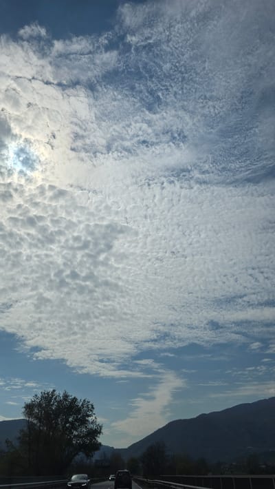 The image shows a sky filled with altocumulus clouds, with the sun partially visible through the clouds. On the right side of the image, a mountain range is visible, along with trees and a highway with cars. The location may be near Cherniche, Bulgaria, based on the mountainous terrain and presence of infrastructure.