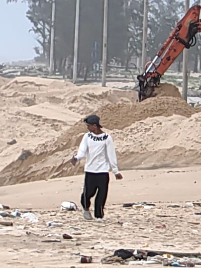 The scene depicts an outdoor sandy area in Thuận An Ward, Vietnam. A single male individual walks from left to right across the foreground. He wears a white long-sleeved shirt with the word "GIVENCHY" printed on the chest, black athletic pants with white side stripes, and a dark baseball cap worn backward. His right hand holds a small, dark, rectangular object. The sandy ground in the foreground is heavily littered with various discarded items, including plastic bottles, wrappers, cans, and other debris. In the mid-ground, numerous uneven sand dunes and piles are visible. To the right, the orange arm of an excavator is partially visible, positioned within a large mound of sand, indicating earth-moving activity. The background consists of a line of tall, slender trees and several vertical poles, all observed under a hazy sky.