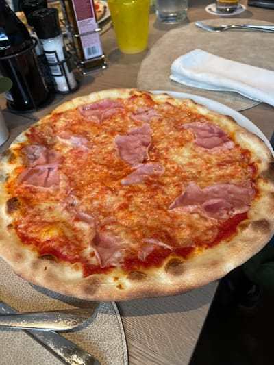 A round, thin-crust pizza with tomato sauce, melted cheese, and thinly sliced ham is presented on a white plate. The pizza sits on a textured placemat on a wooden table. In the background, salt and pepper shakers, a bottle, and a yellow cup are visible. The scene is set in a restaurant in La Ila - Stern - La Villa, Italy.