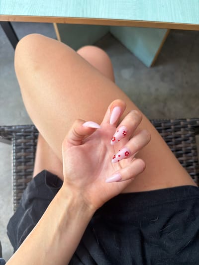 A close-up, overhead shot shows a person's left hand with long, almond-shaped, light pink nails, adorned with small red floral designs on some of the nails. The hand is resting on the person's lap, which is covered by black fabric that appears to have some light-colored fibers on it. The person is wearing black shorts. The background features a woven chair and the edge of a light teal table. The scene is lit by natural light. This image does not provide any information about the city of Prosena, Bulgaria.