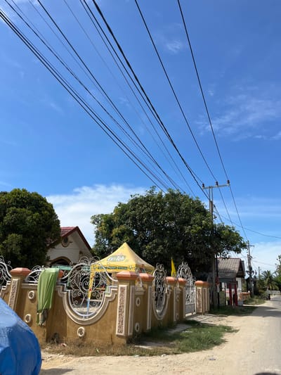 The media file is a photograph taken on a sunny day in Dalam, Indonesia, likely in the late morning or early afternoon given the bright sunlight and clear blue sky. The scene depicts a street with a light pole and numerous overhead electrical wires crisscrossing the frame. In the foreground, there is a decorative wall with ornate metalwork and textured panels, suggesting a residential or commercial property entrance. A yellow awning with visible text can be seen over a section of the wall, partially obscuring a building behind it. A green cloth is draped over an object, and a blue tarp is visible in the lower right corner. Lush green trees are present, adding to the tropical feel of the location. The road or path in the foreground is unpaved, with some grass growing along the edges. The overall impression is of a typical street scene in a warm climate, with infrastructure and architectural elements common in the region. The visible text on the awning reads "Poci Es Teh", which is a brand of iced tea popular in Indonesia.
