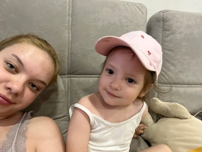 The media file is a selfie-style image taken indoors, likely in a home setting in Gorodets, Russia. Two people are visible, a young child and an adult, both lying down on what appears to be a sofa.
The child, in the foreground and on the left side of the image, is wearing a white tank top and a pink baseball cap adorned with embroidered cherries. The child is looking directly at the camera with a playful expression. To the right of the child is the adult, whose head is also angled towards the camera. The adult is wearing a light-colored, possibly gray, lace-trimmed top. Both individuals are positioned upside down relative to the viewer, suggesting the photo was taken from a low angle or that the camera was held in an unusual orientation.
The background consists of a textured gray sofa. There are no clear indications of the time of day or weather. The scene is intimate and casual, suggesting a relaxed moment between the child and the adult. No visible text is present in the image. The overall mood is one of warmth and familial connection.