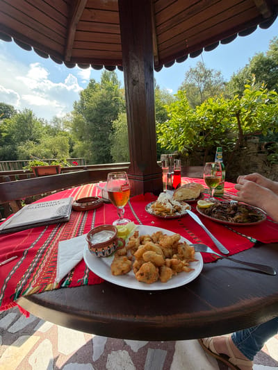 The image displays an outdoor dining scene at "Механа Войводницата" (Mehana Voyvodnitsata), a restaurant located in Zlati Voyvoda, Bulgaria. The perspective is from under a dark brown wooden pavilion or gazebo with a scalloped ceiling edge and a prominent wooden support pillar on the right. Beyond the structure, a bright blue sky with white cumulus clouds is visible above lush green trees and foliage, with a wooden fence discernible in the background. Sunlight indicates a clear, daytime setting, casting shadows on the dark wooden table and the light-colored mosaic-patterned stone floor beneath.
A large, round, dark wooden table is covered with a vibrant red tablecloth featuring traditional Bulgarian woven patterns in red, white, yellow, green, and black. Various food and drink items are arranged on the table for at least two diners. On the left, a large menu titled "МЕХАНА ВОЙВОДНИЦАТА" (Mehana Voyvodnitsata) rests beside a decorative wooden ashtray. A Stella Artois branded goblet, approximately half-filled with amber beer, sits further right.
In the center, a white oval plate holds numerous pieces of deep-fried, battered food, accompanied by a small, patterned ceramic bowl of white sauce and a lemon wedge. To its left, a small oval plate contains french fries or fried potatoes topped with grated white cheese. To its right, another oval plate presents a dark, sautéed dish, possibly mushrooms and peppers, garnished with a lemon wedge. A piece of bread is on a separate small plate.
Beverages include a green Stella Artois beer bottle with Bulgarian text, and two clear bottles, one with red liquid (possibly wine or juice) and another with clear liquid. A second goblet, containing a pale yellow-green liquid, is also present. A set of salt and pepper shakers with red and clear liquids stands near the bread. Cutlery, including a fork and knife, is laid out.
A person's hands and forearm are visible on the right side of the table, holding a fork, indicating they are in the process of eating. Their lower leg and foot, wearing a light-colored sandal, are visible beneath the table. White paper napkins are placed under some plates and near the menu.
