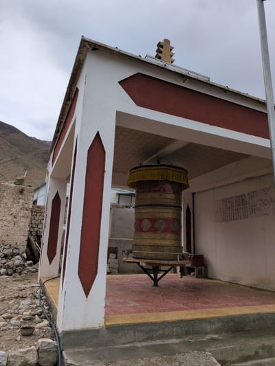 The image depicts a prayer wheel in a covered structure, likely a temple or shrine, in Khardung, India. The structure has white and maroon painted walls and pillars with a tiled floor. A large, ornate prayer wheel, golden with red and yellow accents, stands centrally within the covered area. The surrounding environment shows a rocky, arid landscape with a mountain in the background. A few stone buildings and debris are visible to the left, suggesting a rural or remote setting. The sky is overcast. There are no people visible in the image.