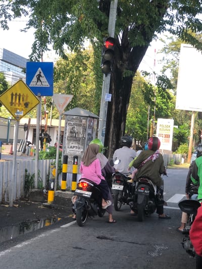 In Banjarkemantren, Indonesia, a street scene unfolds with multiple motorcycles at a traffic light. A traffic light with a red signal is visible. Several individuals are present, primarily riding motorcycles. One motorcycle, with license plate "S 3696 NAY", is prominently featured. Another motorcycle displays license plate "F 6920 FB". On one of the motorcycles, a child wearing a pink jacket is seated between two adults. The environment includes trees with green foliage, traffic signs such as a pedestrian crossing sign and a yellow diamond-shaped sign with vertical lines, and buildings in the background. The road surface is asphalt and appears wet in places, suggesting recent rain. The overall scene depicts a typical urban traffic intersection.
