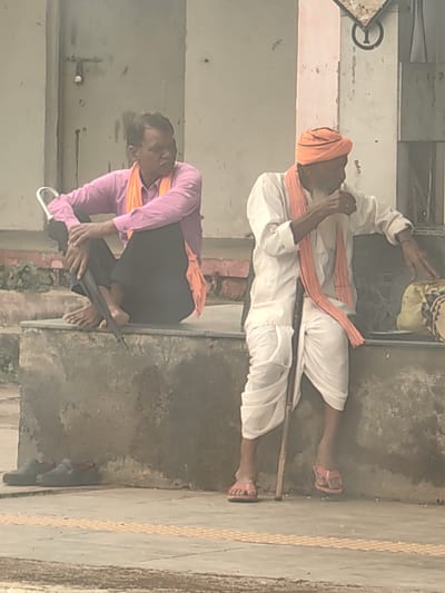 Two men are sitting on a concrete platform. The man on the left is wearing a pink shirt, black pants, and an orange sash. He is holding a black umbrella with a silver handle and has his legs crossed. The man on the right is wearing a white kurta and dhoti, a red turban, and an orange sash. He is holding a walking stick and appears to be eating something. A light-colored bag is to his right. In front of the platform, on the ground, are a pair of dark-colored shoes. The background consists of a light-colored wall with a metal door on the left. The image is taken outdoors, likely in Chhanera, India, given the style of dress and the general environment.