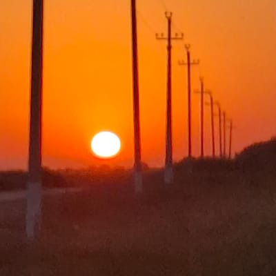 A wide shot captures a vibrant sunset in Қарааспан, Kazakhstan. The sun, a brilliant white orb, is positioned low on the horizon, casting an intense orange glow across the sky. A row of utility poles, silhouetted against the sunset, march across the frame from left to right. The poles are dark and slender, with crossbars visible on some, suggesting they carry electrical wires. In the foreground, the ground is covered in dry grass and low vegetation, rendered in muted browns and reds by the sunset light. The overall atmosphere is one of serene twilight.