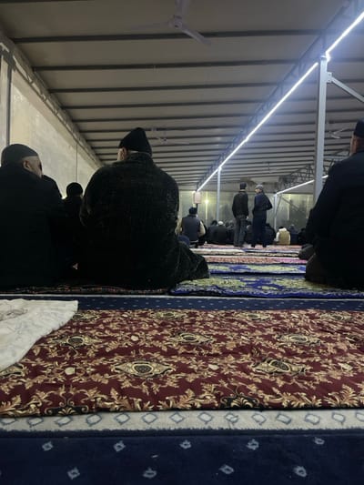 The image presents an interior view of a prayer hall or mosque located in Karmana, Uzbekistan. Approximately 15 to 20 adult males are visible, mostly seated on patterned prayer mats, with their backs to the camera, indicating they are facing a common direction. Most individuals are dressed in dark winter attire, including jackets and hats such as black beanies or kufis. In the immediate foreground, a prominent burgundy prayer mat with gold and black floral patterns is visible, alongside a segment of a white patterned textile. Beyond this, rows of blue and green patterned prayer mats extend across the floor. The ceiling structure features exposed metal beams supporting a light-colored corrugated or panel roof. Multiple white ceiling fans are mounted, and long, linear LED strip lights illuminate the space along the beams. In the mid-ground, two men stand upright, appearing to be in conversation, while others remain seated in various states of repose or prayer. The environment is well-lit and appears to be a functional space for communal gathering.