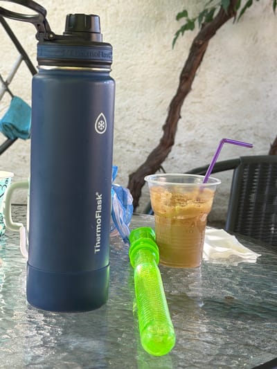 The image depicts a close-up, eye-level view of a glass-topped table outdoors, likely in a garden or patio setting. The focus is on several objects arranged on the table.

On the left is a large, dark blue insulated water bottle, branded "ThermoFlask," with a black lid and a blue band around the neck. Next to it, partially visible, is a light blue mug with a white handle and a pattern of light blue hearts.

In the center, to the right of the water bottle, is a clear plastic cup filled with a frothy, light brown beverage. A vibrant purple straw extends from the drink and curves upward.

In the foreground, a bright green, ribbed plastic object, possibly a bubble wand or a toy, lies horizontally on the table.

In the background, a light-colored textured wall is visible, with a dark, gnarled tree branch extending across the frame. A wicker chair is partially visible in the upper right quadrant. A white paper napkin is placed near the plastic cup.

The lighting suggests it is daytime, with sunlight casting reflections on the wet-looking glass table surface. The scene appears to be set in Krumovgrad, Bulgaria, outdoors on a table, possibly at a cafe or residence. No people are visible in the image.