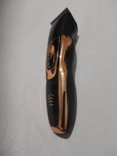 A black and gold electric hair trimmer is positioned vertically on a white, textured surface. The trimmer has a metallic gold accent that runs down the center of its body, contrasting with the matte black casing. At the top of the trimmer, a metal blade is visible. A small, rectangular button is also present on the gold accent. Below the button, a logo or brand name, appearing to be "Geemy", is faintly visible in gold lettering. Further down the trimmer, there are three sets of small, gold chevron-like arrows pointing downwards. The background is a plain white fabric, with subtle shadows cast by the trimmer, indicating it is the sole subject in the frame. No people or specific activities are depicted, and no environmental context such as time of day, weather, or specific location cues are provided beyond the brand name.