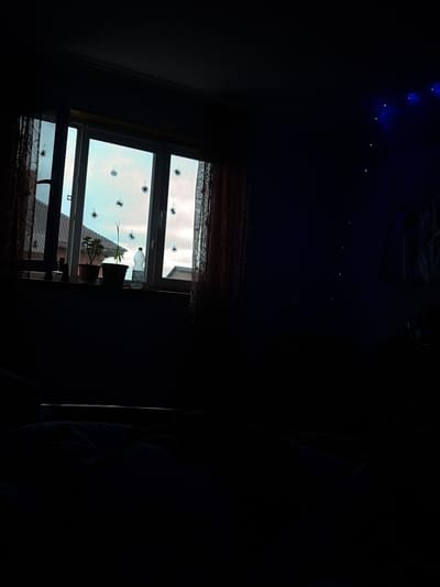 The image depicts an indoor scene, likely a bedroom, at dusk or night. A window with sheer curtains on either side is the primary light source, revealing a pale blue sky with some clouds outside. Several black, star-shaped decorations are adhered to the windowpanes. On the windowsill, there are two potted plants and a plastic bottle. To the right of the window, a string of blue fairy lights is visible against a dark blue wall. The foreground is dominated by darkness, with the impression of a bed covered in blankets. The scene is dark and subdued, with the artificial lights creating a moody atmosphere. The image does not contain any specific visual cues that would definitively place it in the city of Leninogorsk, Russia.