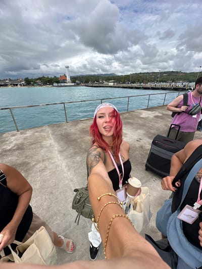 A group of people are on a concrete pier next to a body of water. A fair-skinned person with pink hair, a white lace headband, and tattoos on their arm is holding up a coffee cup and smiling. This person is wearing a black top and a pink lanyard. In the foreground, another person's arm with gold bracelets is visible, as well as a person's leg wearing a tan sandal. The person is also sitting next to an olive-colored purse. Two other people are standing with luggage. In the background, a view of Kampung Toyapakeh, Indonesia is seen, with buildings, a lighthouse, and cloudy skies. The concrete pier has a metal railing.
