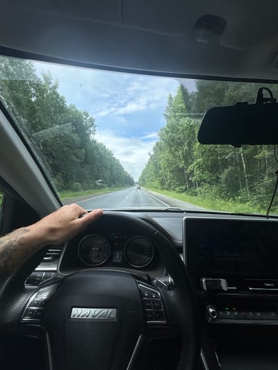 The image is a first-person perspective from inside a Haval car, driving on a paved road bordered by dense green trees. The driver's left hand is on the steering wheel. The dashboard displays a digital speedometer reading 0 km/h and an odometer reading 111265 km. The car is stationary, indicated by the zero speed. The sky is partly cloudy and bright. The scene appears to be a rural or wooded area, possibly on the outskirts of Uspenskii, Russia, given the context of the location. There are no other people clearly visible in the frame. The road ahead shows a slight incline and a few other vehicles in the distance.