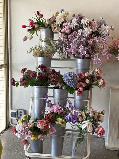 A tiered metal display rack holds an assortment of artificial floral arrangements. The rack is positioned against a textured, off-white wall. To the left, a window with horizontal blinds is partially visible.
The floral arrangements vary in color and type, featuring a spectrum of pinks, purples, whites, and some yellow accents. Many of the flowers are clustered in galvanized metal buckets of various sizes, while others are in glass vases.
Starting from the top shelf, a bucket contains a dense arrangement of flowers in shades of pink and white, with some dark red tulips visible. The second shelf down holds a bouquet with lighter pink and white flowers. The third shelf down features a bucket with clusters of deep red or burgundy flowers. The fourth shelf has a bucket containing a mix of pink and purple flowers, including a prominent blue hydrangea. The bottom shelf includes a bucket with a colorful assortment of pink and white roses, as well as yellow flowers. To the right of this, a glass vase holds a bouquet with lighter pink flowers, purple accents, and white lilies.
In the lower left corner, a gray electrical outlet box with two receptacles is mounted on the wall. The floor appears to be concrete. The overall scene suggests a display, possibly in a shop or a decorative setting, showcasing artificial flowers. The specific location in Tam Kỳ, Vietnam cannot be determined from the visual information provided.