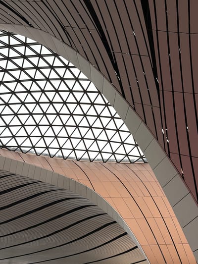 This image showcases the interior architecture of the Beijing Daxing International Airport in Guangyang District, China. The scene is characterized by sweeping curves and geometric patterns formed by the airport's structural elements.

Dominating the upper portion of the image is a large, triangular-grid skylight, revealing a bright, overcast sky. The metallic framework of the skylight creates a tessellated pattern of intersecting lines.

Beneath the skylight, a series of curved architectural surfaces are visible. The main subject is a large, sweeping metallic structure with a warm, copper-toned finish. This surface is composed of many rectangular panels, with dark lines demarcating the seams between them, creating a visually rhythmic effect. This copper-toned surface appears to curve downwards and towards the viewer.

Above and to the right of this copper-toned element, another curved structure is present. This one is clad in a reddish-brown material, divided into vertical, slightly curved slats. These slats are separated by dark gaps, giving the impression of a ribbed or fluted surface. This element also appears to curve and recede into the background.

Further below and to the left, another layered architectural element is visible. This surface is also curved and features a pattern of parallel, dark, wavy lines against a lighter background, creating a sense of depth and undulation. This element appears to be beneath the copper-toned structure.

The lighting is diffuse and even, suggesting an interior space illuminated by natural light from the skylight and possibly artificial sources not directly visible. There are no people or any other objects or elements that would indicate activity or specific purpose within the frame. The overall impression is one of modern, aerodynamic design with a focus on flowing lines and geometric repetition.