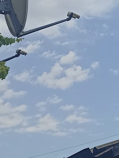 The image depicts an outdoor scene with a bright, clear blue sky filled with scattered white clouds. In the foreground, the edge of a dark, tiled roof is visible at the bottom of the frame. Extending upwards from the lower right corner, a thin dark line, likely a power line, stretches across the sky.
Dominating the upper left quadrant of the image is a large, dark satellite dish, angled downwards and to the right. A dark pole extends from the dish, supporting it. Attached to this pole, and further down to the left, is a smaller pole with a white and black camera mounted on its end. Another similar pole with a camera is visible extending from beneath the satellite dish, further to the left and slightly lower. Green foliage, possibly from a vine, is also visible in the lower left, partially obscuring the lower camera. The overall lighting suggests it is daytime, likely late morning or early afternoon, with good visibility. The setting appears to be an outdoor location, possibly a residential or commercial area given the presence of the satellite dish and cameras. No people are visible in the frame. There is no discernible text in the image. The overall mood is neutral and observational.