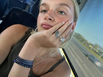 A young, fair-skinned female, with blonde hair and green eyes, is depicted seated inside a vehicle, presented from the chest up. She wears a small nose ring in her right nostril, large silver hoop earrings, and a dark olive or grey sleeveless top with a bronze-colored D-ring detail on the shoulder strap. Her right hand rests against the right side of her face, with her fingers slightly curled. On her right ring finger, she wears two silver rings: one with an intricate, branching design, and another featuring an oval, light-colored cabochon. Her left wrist displays multiple dark blue bracelets, including several leather-like wraps and a strand of beaded stones. Two delicate silver chain necklaces are visible on her neck. Behind her to the left, a dark blue vehicle seat with some texture is partially visible. To her right, a window reveals a blurred exterior view of a multi-lane highway with distant vehicles, green foliage, and a clear blue sky, all partially obscured by horizontal lines on the glass. The scene is situated within a moving vehicle in the city of Whitby, Canada, as specified.