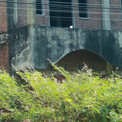 The image shows a brick building with a weathered, concrete facade and several horizontal wires running across the upper portion. In the foreground, there is lush green foliage with small pink flowers, obscuring the lower part of the building. The building appears to be in a state of disrepair, with peeling paint and exposed rebar on the lower concrete section. An arched opening made of brick is visible in the central part of the facade, with small rectangular windows or openings within it. The overall scene suggests an outdoor environment in a tropical or subtropical climate, given the dense vegetation. The context of Keutapang, Indonesia, implies a location that experiences such conditions.