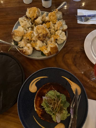 The image displays a dining scene from an overhead perspective, set on a polished dark wooden table, consistent with a restaurant environment. The exact location within Santiago, Mexico, cannot be determined from the visual information provided.

In the upper left quadrant, a light-colored plate contains a substantial portion of roasted or fried cauliflower florets, many of which are generously coated with an orange-colored sauce. A silver fork is inserted into the cauliflower, suggesting it is being served or has been partially consumed.

In the lower right quadrant, a dark blue ceramic plate holds a circular, dark-brown griddled item, possibly a cheese disc or sopes, topped with a mound of green, leafy ingredients, likely avocado or lettuce, garnished with thin, crispy, golden-brown strands (possibly fried onions or tortilla strips). Three decorative swirls of a light orange-beige sauce are present on the plate's rim. A knife and fork with dark handles are positioned to the right of this dish.

Additional elements include a folded white napkin with cutlery and a white ceramic plate in the upper right. To the mid-right, the base of a stemmed glass is visible, with a small amount of red liquid at its bottom. A section of a black, possibly leather, object is visible on the far left. Two blurred, white cylindrical objects, likely condiment shakers, are in the background. No individuals are present in the frame.