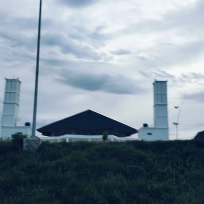 A wide, slightly blurry shot captures a modern mosque building and its surroundings under an overcast sky in Cotkareung, Indonesia. The central structure of the mosque features a dark, steeply sloped, pyramidal roof, with a lighter-colored base visible underneath.
Flanking the main building are two tall, white, rectangular minarets. A small, blue water tank is visible behind the base of the left minaret. Near the right minaret, thin, linear structures, possibly antennas or power lines, ascend towards the sky. A tall, dark, slender pole runs vertically through the left side of the frame, extending from the bottom to beyond the top edge.
The sky is filled with soft, light grey and white clouds, indicating an overcast or hazy day. The lighting is muted, contributing to a tranquil and somewhat subdued atmosphere. The foreground consists of a dense field of dark green grass, which appears blurred, suggesting either motion or a shallow depth of field. Small, dark bushes are also visible near the base of the mosque and minarets.
No people are visible, and there is no specific activity taking place. No text is discernible within the image.