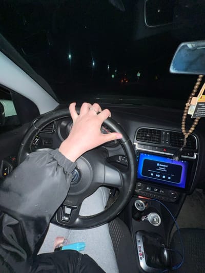 Inside a car, a person's hand rests on the steering wheel, obscuring the VW logo. The dashboard displays a modern infotainment system with music controls and Bluetooth connectivity. Two partially visible aluminum cans sit near the gear shift. The passenger seat has a glimpse of gray fabric and a blue pacifier. Outside, the night is dark with blurry lights, as seen through the windshield and side mirror.  A string of wooden beads and a hanging tag dangle from the rearview mirror.