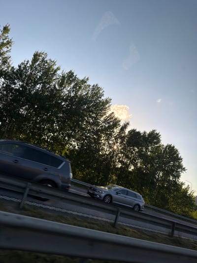 The image captures a view from a moving vehicle on a highway, likely in or near Kungälv, Sweden, given the context. The scene is dominated by a clear blue sky with a few wispy clouds. Sunlight filters through the dense green foliage of trees lining the highway, creating dappled light and shadow. Two cars are visible on the road; a dark-colored SUV in the foreground on the left lane, and a silver station wagon further ahead in the right lane. The cars are in motion, indicated by a slight blur. The highway itself has multiple lanes delineated by white lines. Metal guardrails separate the lanes and the roadside. The overall impression is one of a late afternoon or early evening drive during a clear day.