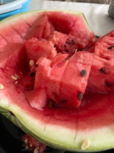 This close-up, overhead shot captures a halved watermelon, its vibrant red flesh bursting with juicy segments. The watermelon is presented in a way that suggests it has been recently cut and is ready for consumption. Several triangular pieces of watermelon are pulled out from the main body, exposing the seeds scattered within the sweet, pink flesh. The rind, a gradient of green and white, frames the fruit.
The setting appears to be an outdoor picnic or a casual meal, possibly in a rural or natural environment, hinted at by the soft, natural lighting and a blurred background that might include outdoor elements. The focus is entirely on the refreshing fruit, with a fork partially visible on the right side, indicating that someone is about to eat it. The image evokes a sense of summer refreshment and simple enjoyment. The time of day is likely daytime, given the bright, even lighting. There is no visible text in the image. The presence of a fork and the way the watermelon is cut suggest a context of a meal being served or prepared. The detail of the cut pieces, some showing the fibrous texture of the flesh, adds to the realism and appeal of the fruit.