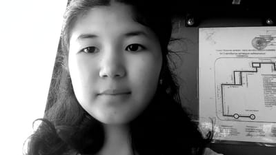 A black and white, medium close-up photograph shows a young woman's face, turned slightly towards the camera, dominating the left and center of the frame. Her dark, wavy hair frames her face. She has dark eyes, a small nose, and her lips are slightly parted. She is wearing a small, rectangular earring in her visible right ear. Behind her, on the right side of the image, a diagram is mounted to a dark surface. The diagram appears to be a map or route plan, with text in a non-English alphabet and numbers. The text above the diagram reads "Проект боюнча келеген жаңы автобустук каттамдын кыймылынын СХЕMASЫ". Below that, it states "№13 автобустук каттамдын кыймылынын СХЕMASЫ". This translates to "Project for the new bus route's movement SCHEMES" and "№13 bus route's movement SCHEMES". A stamp with Cyrillic text is visible in the upper right corner of the diagram, which appears to be related to a governmental or official entity. The overall environment appears to be indoors, possibly a bus station or administrative building in Жаны-Кыштак, Kyrgyzstan, given the context of a bus route diagram.