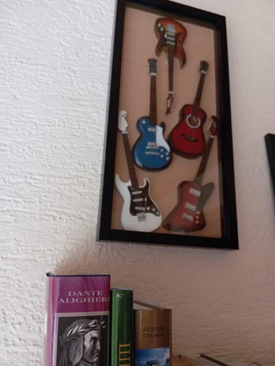 The image shows a close-up of a framed collection of miniature guitars hanging on a textured white wall. The frame is black and contains at least five guitars of various colors, including red, blue, and white. Below the framed guitars, a portion of a bookshelf is visible, holding several books. The leftmost book is purple with the name "DANTE ALIGHIERI" printed on its spine, along with an image of a man with a laurel wreath. To the right of this book, a green book is visible, followed by a brown book with the name "AEDDEN CREMIN" printed on its spine. The scene appears to be an indoor setting, possibly a home or a small shop, given the decorative display and the presence of books. There is no indication of the city of Baden, Austria, in the image.