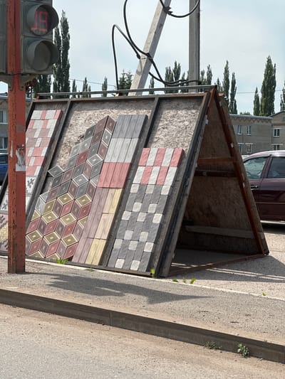 An outdoor display of paving stones is set up near a traffic light in Novaya Otradovka, Russia. The paving stones are leaning against an A-frame structure made of wood and metal. The display features various patterns and colors of paving stones, including red and white checkered patterns, diamond shapes in red, yellow, and grey, and wood-grain textured stones in pinkish-tan and grey. In the background, there are tall, slender trees, brick buildings, and a dark red car. A traffic light showing a red "19" is visible on the left side of the image. The ground is paved with asphalt and gravel, with a concrete curb. The scene is illuminated by bright sunlight, casting shadows on the ground.