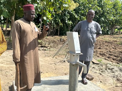 In Afogo, Nigeria, two men stand near a hand pump in a rural setting. The man on the left, wearing a brown tunic and a maroon cap, holds a green mobile phone. The man on the right, in a grey tunic, stands with his hands behind his back. The hand pump is a metallic structure with a long handle. The ground is dry earth, with scattered vegetation and a backdrop of lush green trees. The scene appears to be under bright daylight, suggesting an outdoor location.