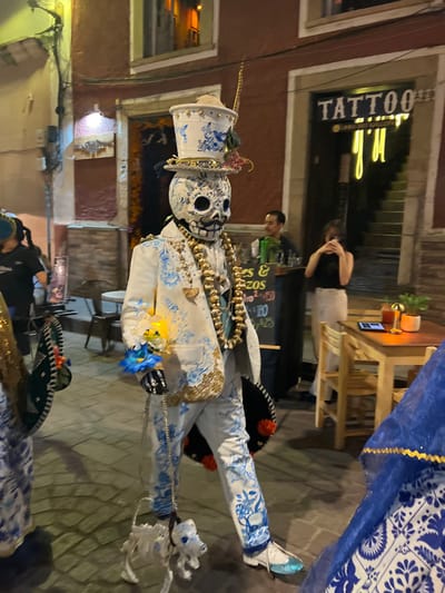 The image depicts a nighttime street scene in Guanajuato City, Mexico. The foreground is dominated by a person in an elaborate skeletal costume, consistent with a Catrín figure for Día de Muertos. This central figure wears a white skull mask with black facial features, a tall white top hat adorned with blue floral patterns, gold trim, a feather, and small decorative elements. The costume includes a white suit with extensive blue floral patterns and gold accents on the lapels, cuffs, and pant legs. Multiple strands of large, gold-colored bead necklaces are worn, and a black sombrero with red poms is attached to the left hip. The figure holds a small bouquet of artificial blue and yellow flowers in the right hand and a silver chain leash in the left, which is attached to a small, white skeletal dog figure with blue accents and a red collar. The figure's shoes are white with metallic blue pointed tips.

To the immediate left, a second costumed individual is partially visible, showing a blue and white patterned garment and a green, fan-like accessory with gold trim. Both costumed figures appear to be walking forward, suggesting a procession or parade.

The background features a narrow street or alleyway lined with buildings with reddish-brown facades. On the right, a business entrance is visible with a "TATTOO" sign and "Urbano Art Gallery" below it, featuring a distinctive neon "J'LL" or similar glowing sign inside. Adjacent to this, an outdoor dining area is present with two wooden tables and chairs. One table holds a smartphone, a small potted plant, and a drink. Behind this area, a male individual, possibly a server, stands near a dark counter or bar, holding a tray with green bottles. A chalkboard sign nearby displays text including "Es & RZOS" and prices "180" and "25." A female individual, wearing a dark top and light pants, stands near the outdoor tables, holding a smartphone up as if taking a photograph. The street is paved with flagstones or cobblestones, illuminated by overhead and building lights.
