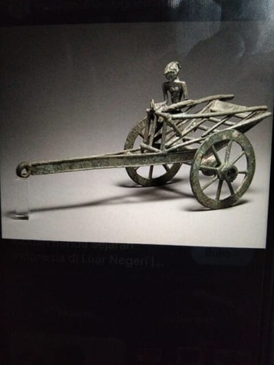 A bronze miniature chariot with a figure is displayed against a plain, light grey background. The chariot has two spoked wheels, a long rectangular body, and a shaft extending forward with a hook at the end. A small, crudely formed figure of a person is seated at the front of the chariot, holding onto the structure. The chariot appears old and weathered, with a green patina on the bronze. There is a shadow cast by the chariot on the surface it rests on. Text is visible at the bottom of the image, indicating it is part of a collection of historical artifacts, possibly related to Indonesia. The specific location within Kualaraja, Indonesia, is not discernible from the image.