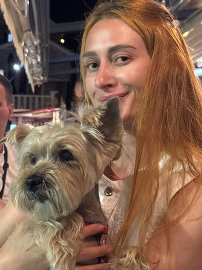 A medium shot captures a young woman with long, reddish-blonde hair holding a small, tan-colored dog close to her face. The woman is smiling, and her head is tilted slightly. The dog, with its fur covering its eyes, appears to be looking forward. They are positioned in front of an out-of-focus background suggesting an outdoor evening setting, possibly a restaurant or cafe, with soft lighting and hints of structures. The scene takes place in Medinski Krš, Montenegro.