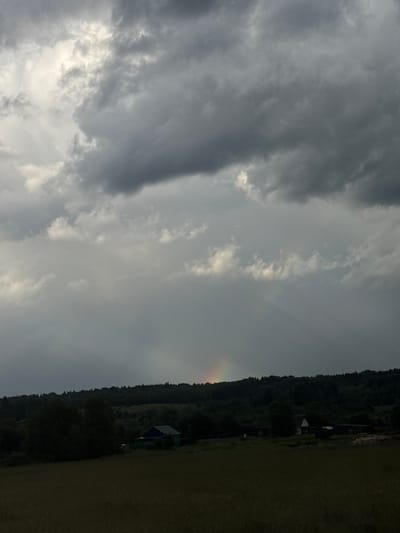 The image captures a rural landscape under a dramatic, overcast sky. Dark, heavy clouds dominate the upper portion of the frame, with patches of lighter sky suggesting a break in the weather. A faint rainbow arcs across the middle ground, its colors muted by the prevailing gloom. Below the sky, a dense line of dark green trees forms a backdrop to a scattered collection of rural buildings. One building on the right appears to be a small, dark-colored house. The foreground is a field of dark, textured vegetation. The overall atmosphere is somber and moody, evoking a sense of stillness. The image is identified as being taken in the city of Laiga, Russia.