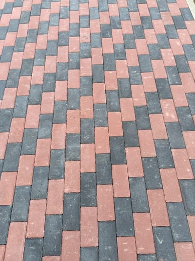An outdoor, ground-level shot captures a section of a paved walkway composed of interlocking rectangular pavers. The pavers are arranged in a repeating pattern of alternating colors: a reddish-brown and a dark gray. The perspective is from a slightly elevated angle, looking down and across the walkway, revealing the texture and spacing of the individual bricks. The scene is devoid of any people or other subjects, focusing solely on the architectural element of the paving. The background is not visible, implying the walkway is part of a larger, unseen urban or suburban environment. This pavement is located in the city of Karkaraly, Kazakhstan.