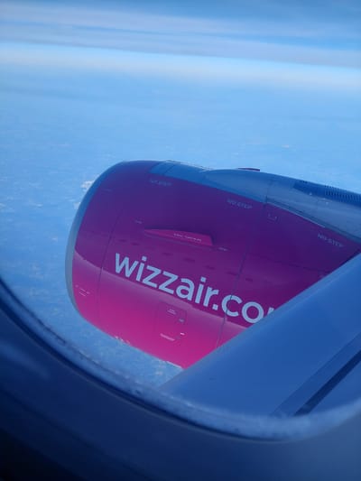 The image, taken from inside an airplane, shows the wing and engine of a Wizzair aircraft mid-flight, with a blue sky and landscape visible outside the window. The engine is painted pink with the white "wizzair.com" logo. "NO STEP" is printed in black on the upper surfaces of the engine. The landscape below is blurry but appears to show terrain. There is no other identifying information about the location of Pyrzowice, Poland. There are no people visible.