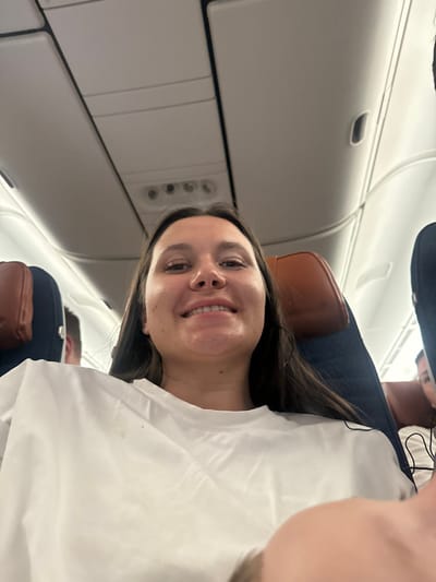 The image depicts a young woman posing for a selfie or being photographed inside an airplane cabin. She is positioned prominently in the foreground, looking directly at the camera with a wide, joyful smile. Her long, dark brown hair is visible around her face, and her dark eyes are bright. She is wearing a simple white top, and a white blanket or sheet is draped over her, covering her chest and left shoulder. Her head appears to be resting against a seat or pillow.
The background reveals the interior of an airplane. Above and to the left of the woman's head, white overhead compartments are visible, along with a panel featuring air vents and passenger service lights. Below and to the right of the woman, another passenger is partially visible, appearing to be a male. This individual is wearing a dark blue garment, possibly a shirt or jacket, and is slumped back in their seat, seemingly resting or sleeping. A brown headrest, possibly leather or faux leather, is visible behind both the main subject and the other passenger. A thin, dark wire, possibly from headphones, can be seen near the second passenger's shoulder.
The lighting within the cabin is artificial, contributing to a slightly dim but well-lit appearance around the main subject. The overall atmosphere conveyed is one of relaxation and contentment, largely due to the woman's cheerful expression. No discernible text is visible in the image.