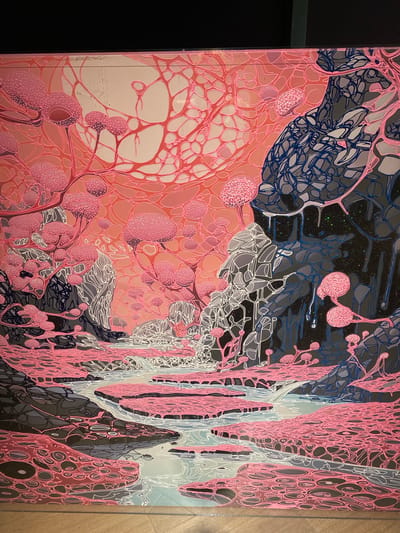 The image is a photograph of a painting that depicts an abstract, alien landscape rendered in vibrant pinks, grays, and blues. The artwork features organic, flowing lines and bulbous, mushroom-like flora. A body of water, possibly a river or stream, winds through the scene, with flat, pink-bordered landmasses on either side. The background is dominated by a large, luminous orb, suggesting a sun or moon, surrounded by a network of delicate pink tendrils. Rocky formations, outlined in white and filled with gray and blue tones, rise from the landscape. There are no people or animals visible in the artwork. The photograph is taken from a slightly angled perspective, showing the painting mounted on a wall. The lighting is even, highlighting the textures and colors of the artwork. There is no identifiable location in Jinniu District, China, within this image, as it is a representation of an imagined environment.